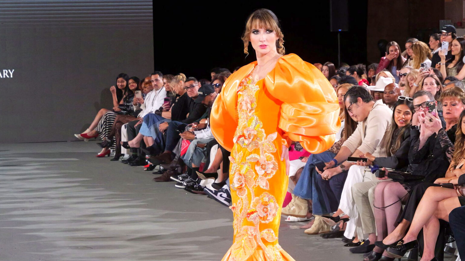 A model in a voluminous orange gown with puffed sleeves walks the runway past a packed front row at a Los Angeles fashion show.