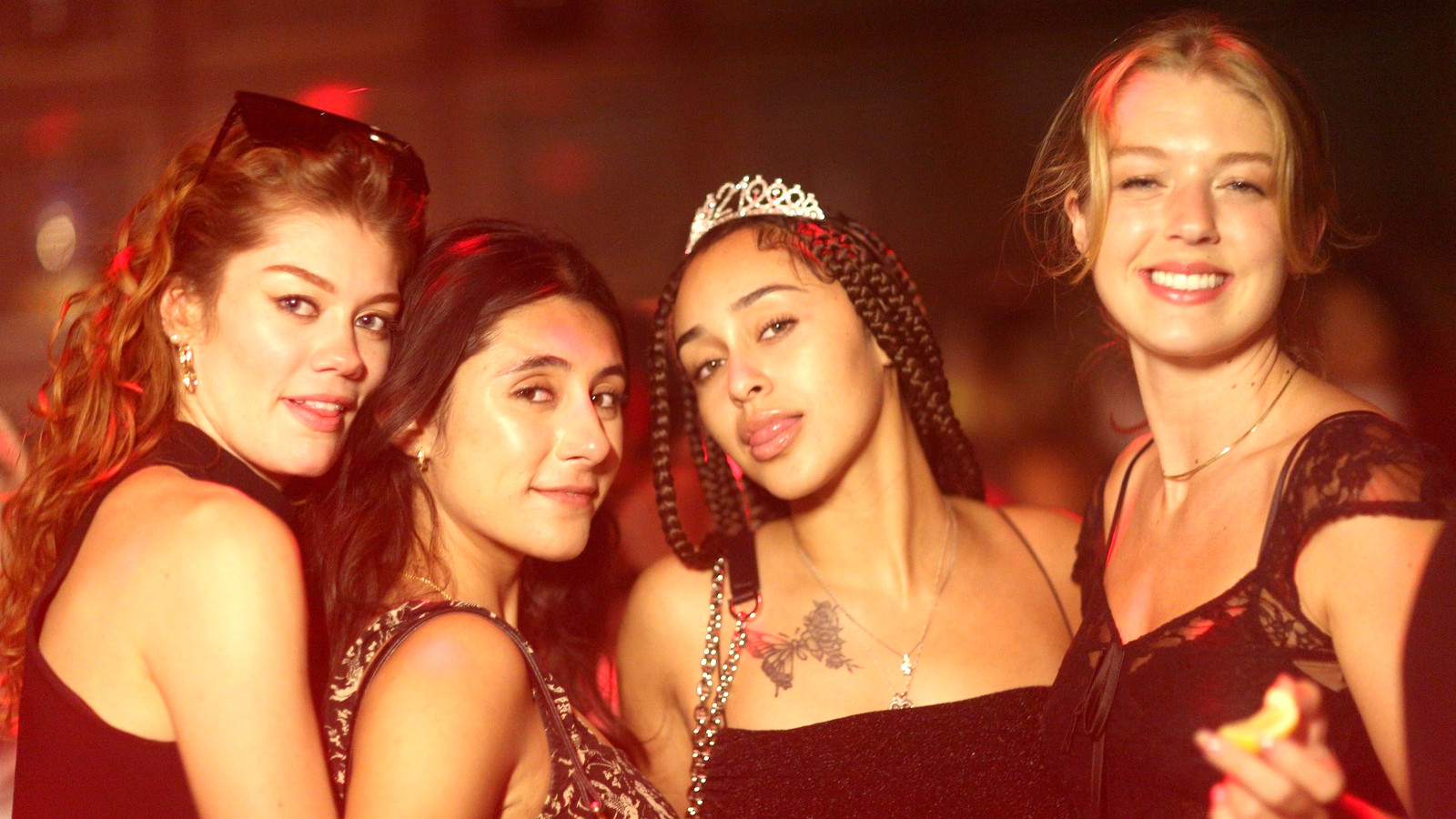 Four friends smile together for the camera in a warm, red-lit bar, one wearing a sparkling tiara.