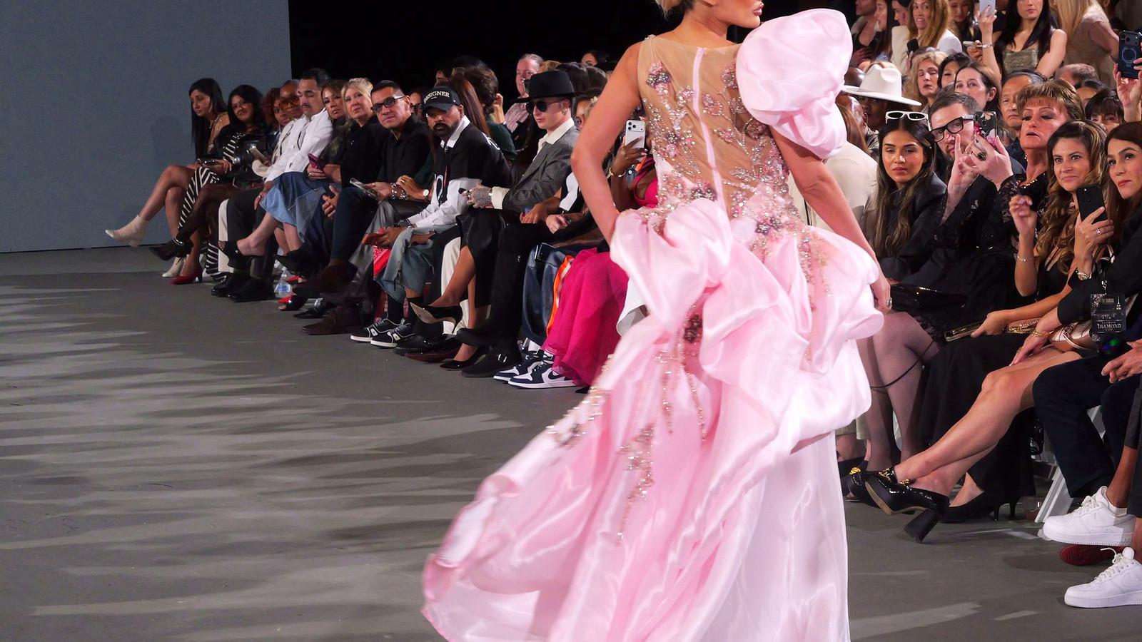 A model in a pink ruffled gown with a sculpted shoulder walks away down the runway as the audience watches from both sides.