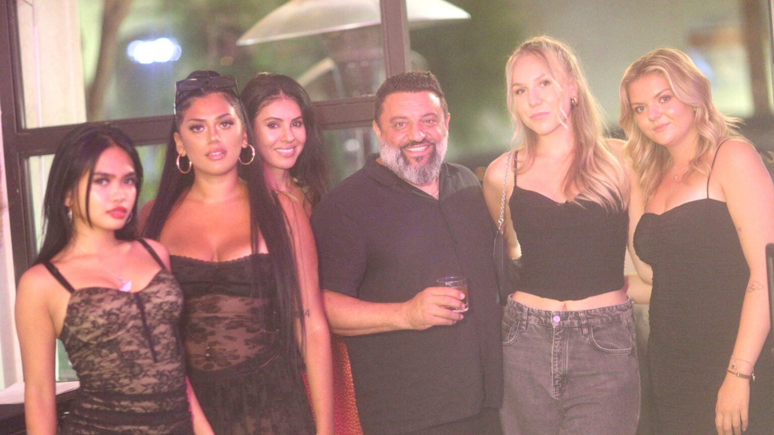A group of friends stands together on a lounge patio under warm string lights, smiling for the camera.