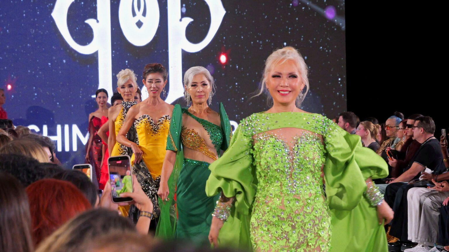 Models in green, gold and red gowns walk the runway together in the show's finale as the audience films on their phones.
