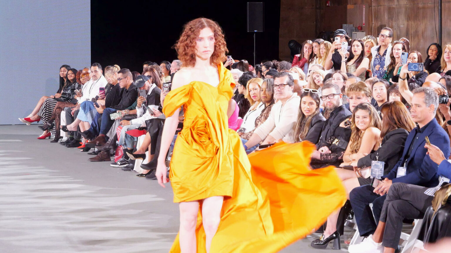 A model in a bright yellow gown with a flowing train walks the runway past a crowded front row.