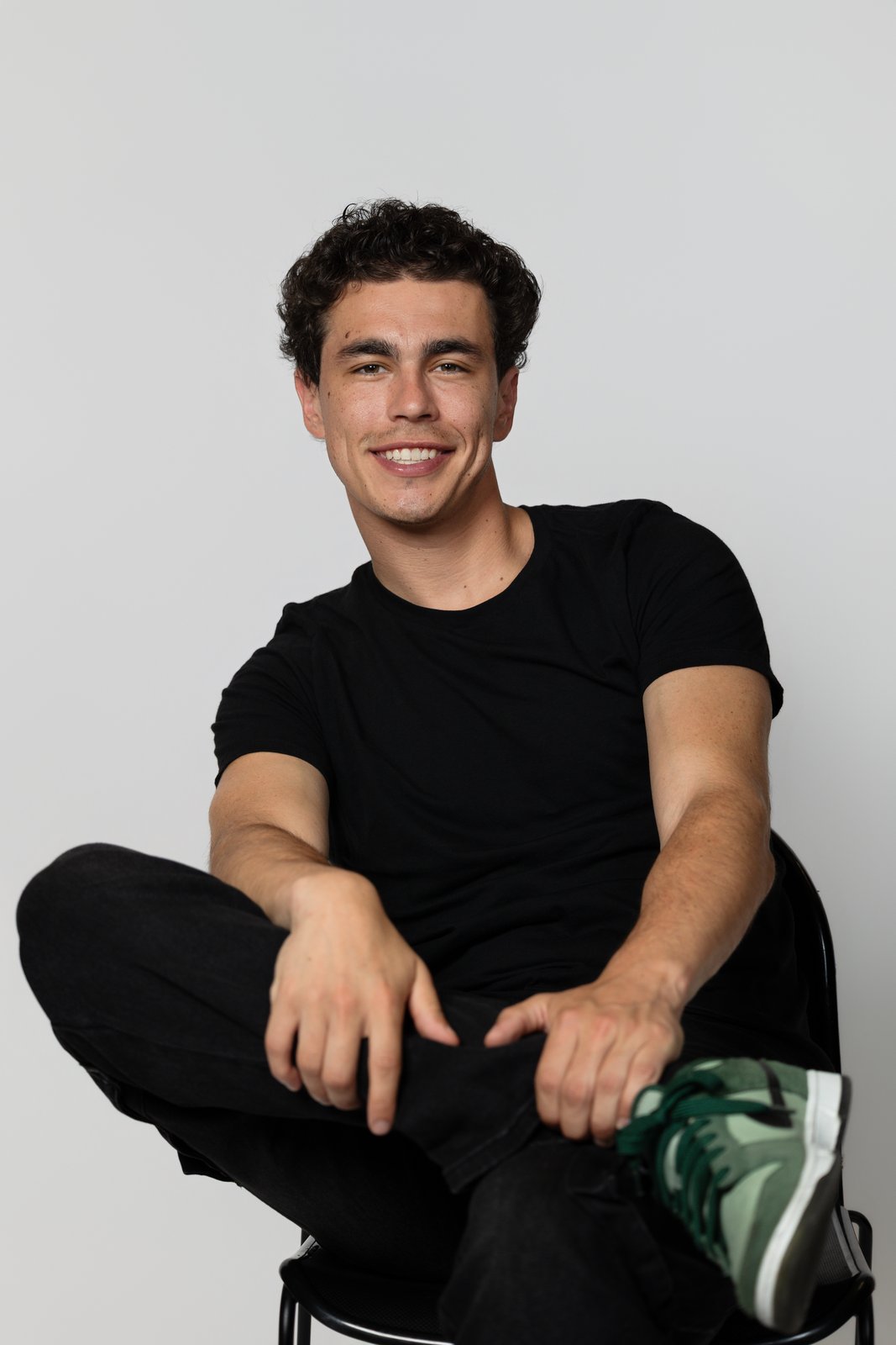 Kevin Fernandez seated on a stool in a black T-shirt and green sneakers against a white studio wall