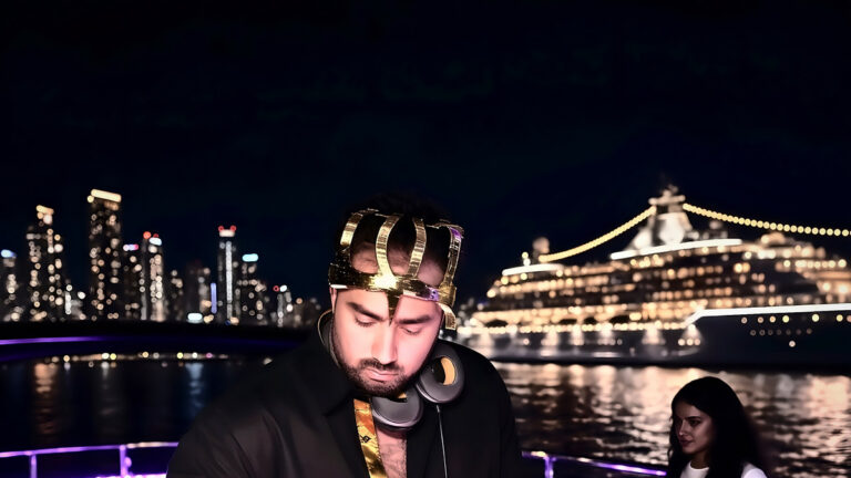 DJ in a gold crown on the top deck of a yacht at night, Long Beach skyline and a lit cruise ship behind